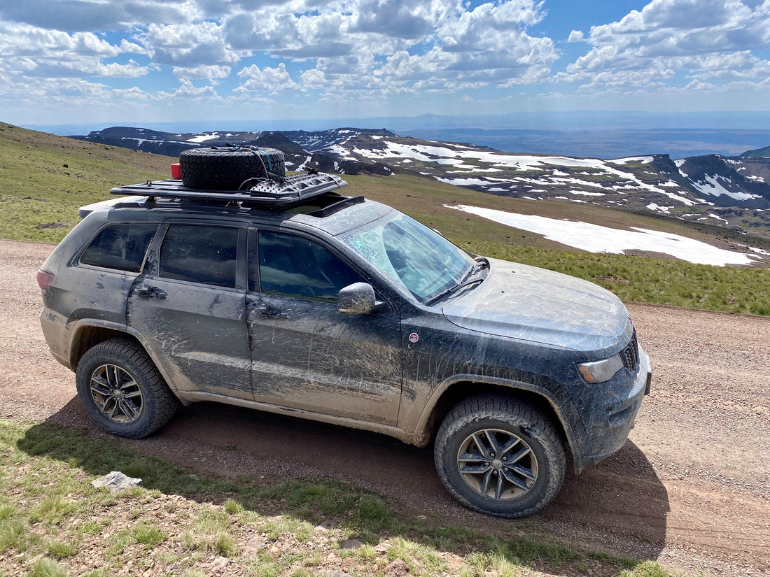 Overland rig, covered in mud, driving on a cliffside trail with beautiful alpine scenery.